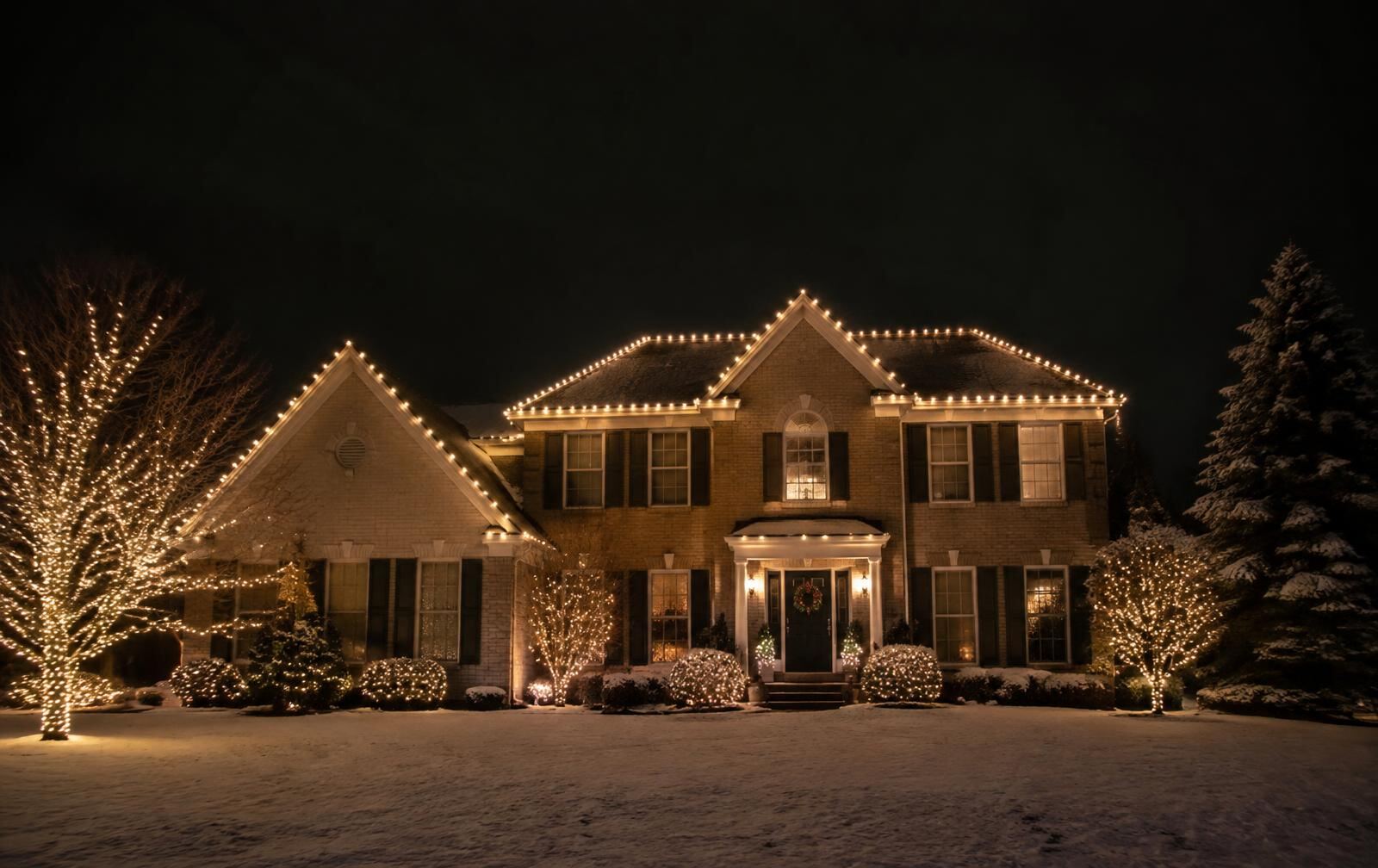 Two-story Northeast Ohio home with warm white Christmas lights on every roofline and wrapped trees at night